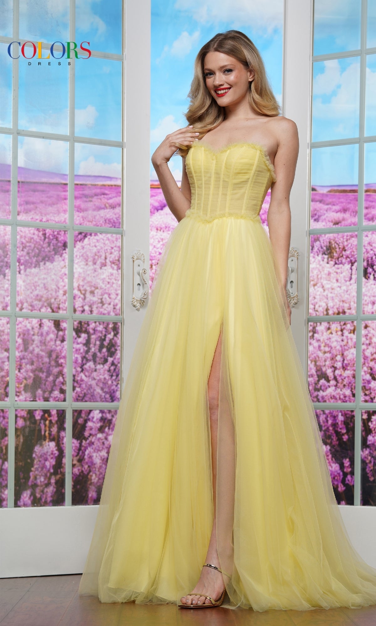 A woman wearing the Strapless Long Prom Ball Gown: Colors Dress 3486 stands indoors before glass doors, a field of purple flowers visible outside.